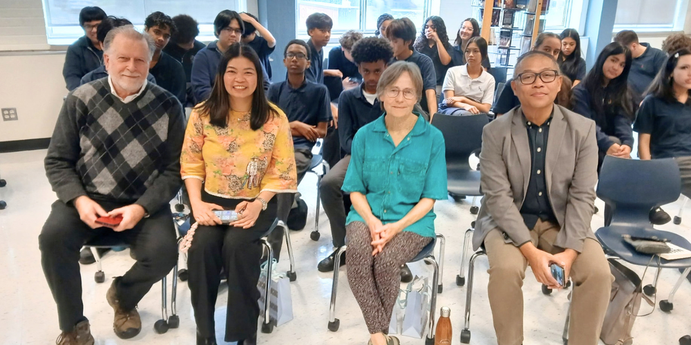 4 adults on chairs, with students in uniform sitting behind them.