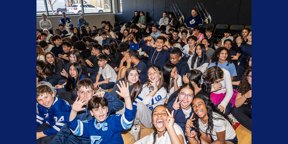 Children sitting on the floor of a gym wearing hockey jerseys.