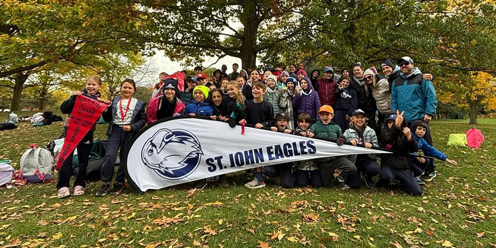 Group photo of St. John students and staff, holding up a sports banner saying "St. John Eagles" with a crest showing an eagle head, and their 2025 TDCAA championship banner