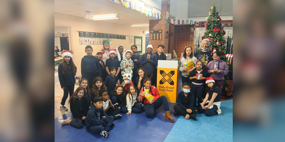 A group of kids and faculty with a Christmas tree and "Daily Bread Food Bank" sign
