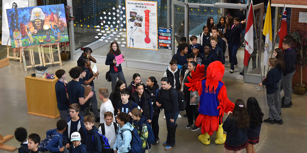 Photo of elementary students waiting in line to get into the festival, while Redmond students hold signs to welcome and guide them, along with a mascot in red bird costume