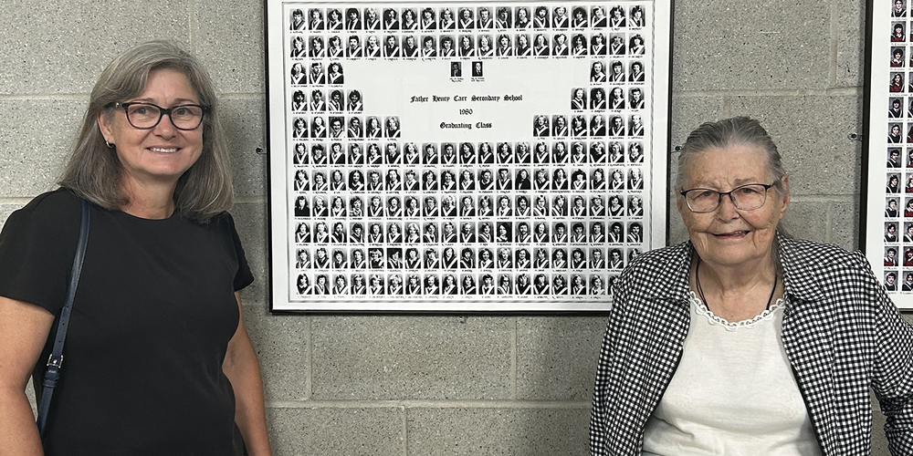 Photo of Deb Smith and Olga Hilmer standing in front of the graduating class plaque that has Father Hilmer's graduating photo.