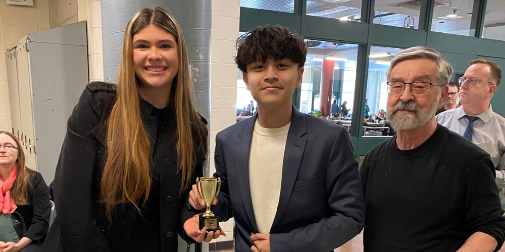 Photo of two students holding an award trophy accompanied by one of the staff