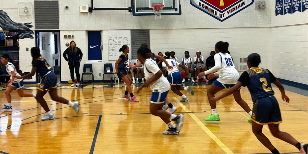 Indoor basketball game with players in blue and white driving toward the hoop, one player in the foreground dribbling on a polished court; gym wall shows banners, a crest logo, and posters.