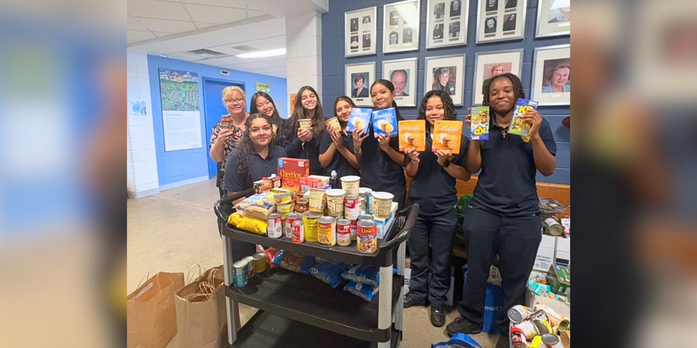 A group of students stand behind a table filled with food and pantry items, smiling and holding up colorful boxes and packages in a school hallway with framed portraits on the wall.