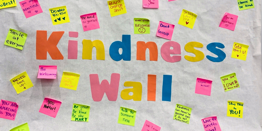 Colorful sticky notes surround the large words “Kindness Wall,” with many small handwritten messages scattered across a light background.