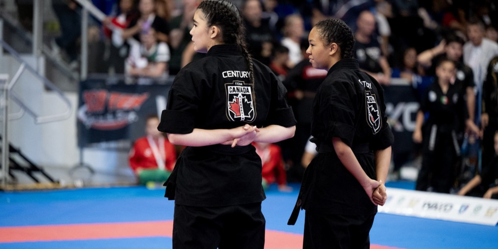 Two martial artists in black uniforms walk on a blue-and-red taekwondo mat during a match, with spectators seated in the background.