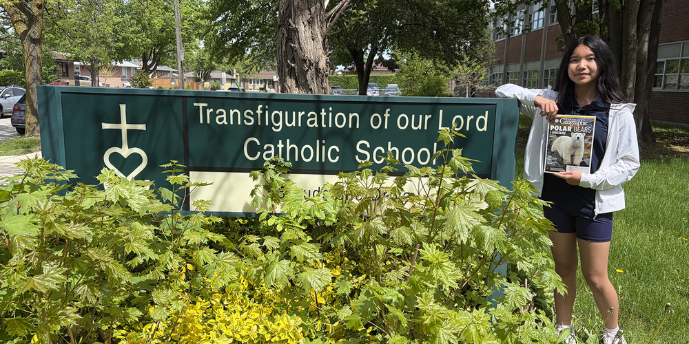Photo of Sinead holding up a Canadian Geographic magazine next to the Transfiguration of our Lord school sign in front of the school building