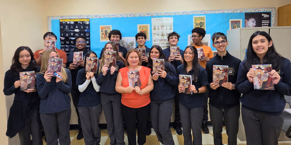 A group of students stand in a classroom holding up printed books or posters, posing for a group photo in front of a bulletin board with colorful displays.