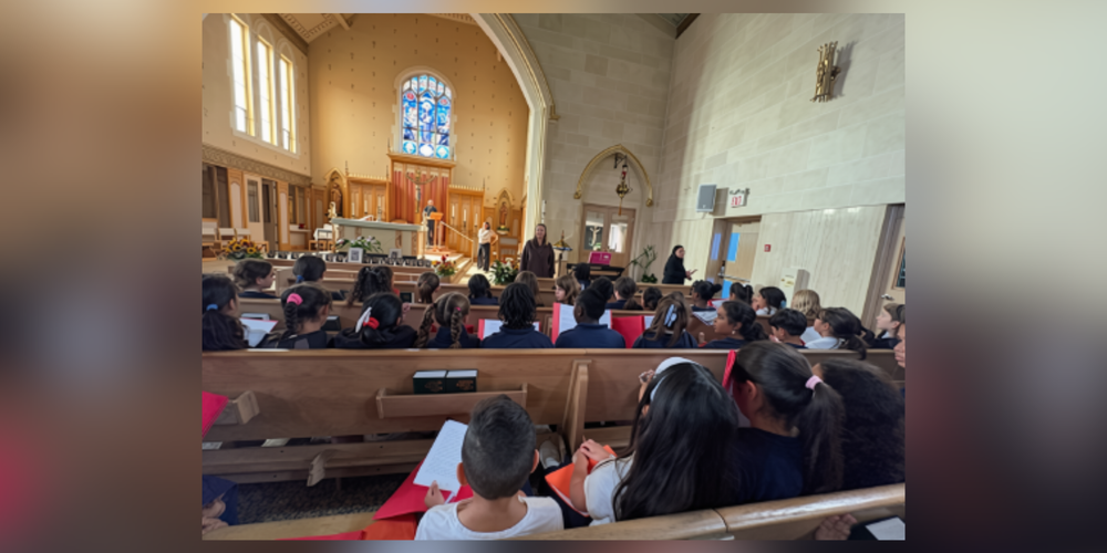Children seated on church pews facing the front of a stone sanctuary, where an adult stands near the altar with a stained-glass window overhead.