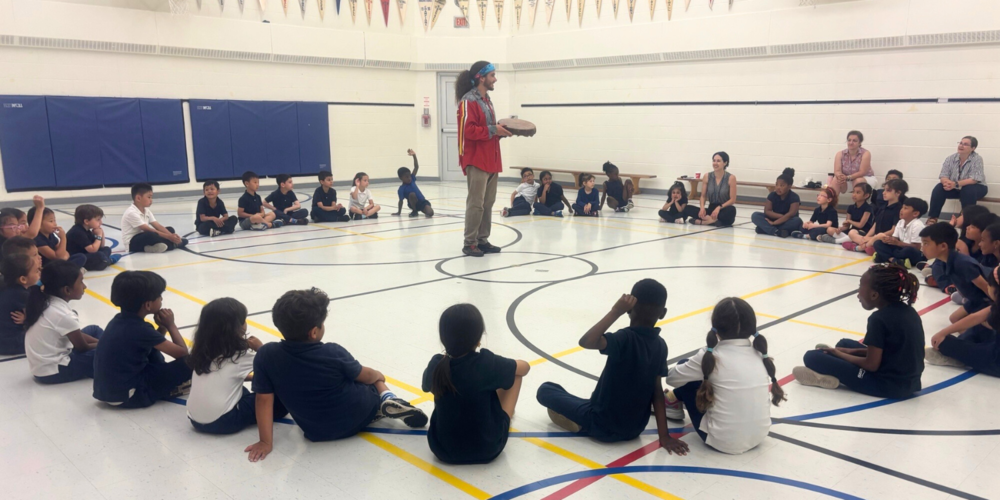 A large group of small kids in a gym, sitting around in a circle around one teacher in the middle who is holding a drum. 