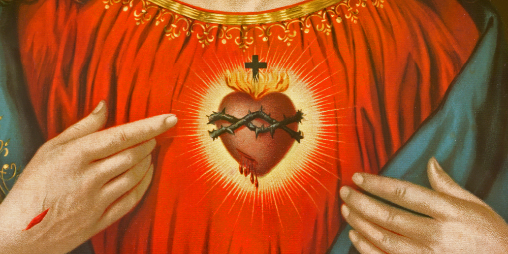 Close-up religious illustration of the Sacred Heart, showing a glowing heart with a cross and crown of thorns at the centre of a red garment. Two hands gesture toward the heart, one with a visible wound, surrounded by radiant light.