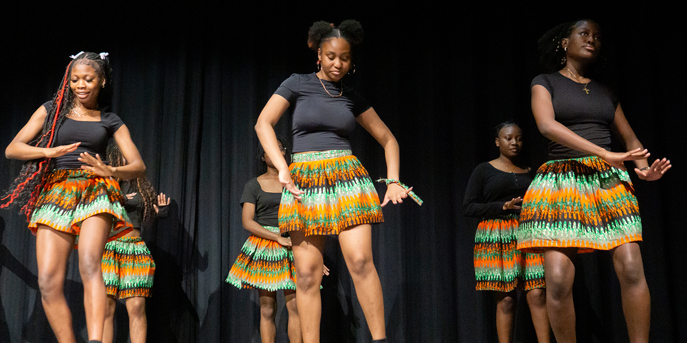 Photo of MPJ students performing a dance on stage while wearing colourful skirts with traditional African patterns