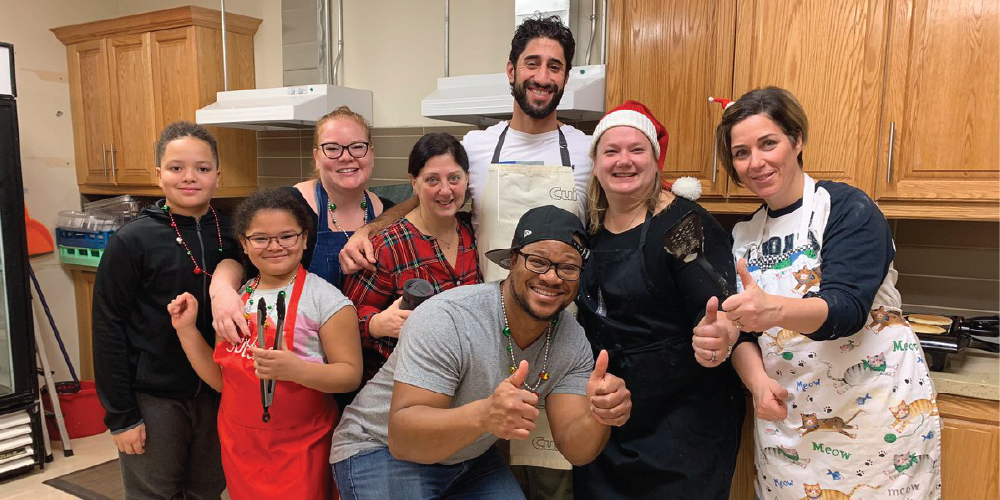 A group of eight people standing together in a kitchen setting, some wearing aprons and festive accessories, posing and giving thumbs‑up gestures. A red banner across the bottom displays the text ‘THANK YOU FOR VOLUNTEERING!’
