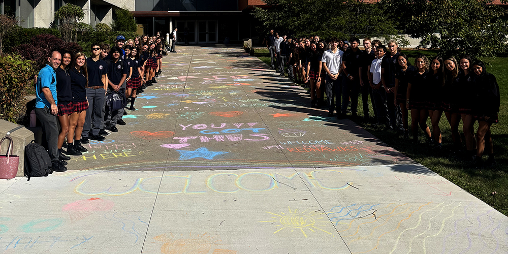 Photo of FJR students and staff in front of the school building with the welcoming chalk messages scrawled on the pavement