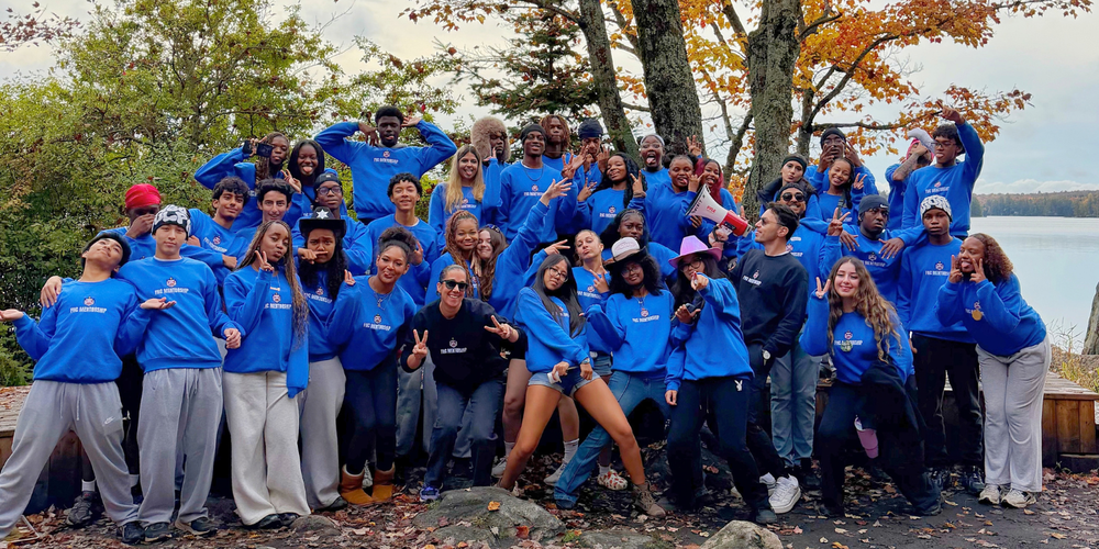 Large group of people in matching blue shirts posing outdoors on a rocky ledge by a lake, with autumn trees and a cloudy sky in the background.
