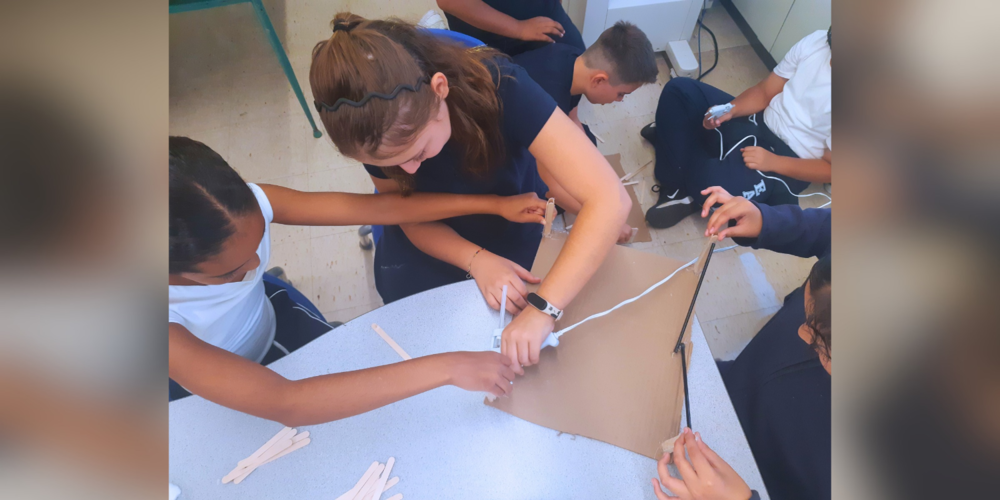 A group of students gathered around a table connecting white wires and components, working together on a hands-on electronics or engineering activity.