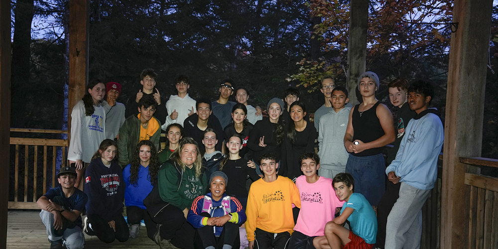 Group photo of Redmond students and staff posing together in their lodge at Camp Muskoka in twilight, with the trees behind them