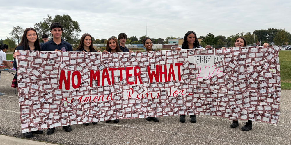 Group of students standing on a paved walkway holding large poster banners covered in red-and-white squares, with bold text “NO MATTER WHAT” and smaller cursive writing beneath.