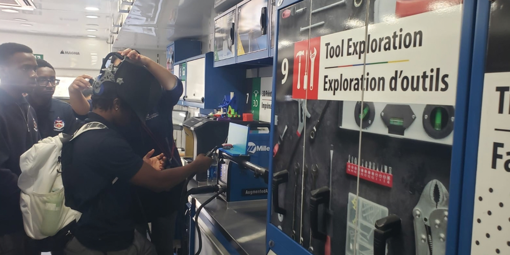 In a workshop, people in the background watch as a blue display case labeled “Tool Exploration” shows tools and gauges, with a person using a device near the equipment.
