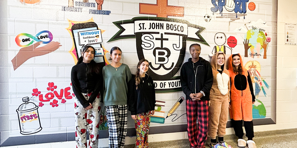Photo of St. John Bosco students in comfy clothing in front of a school wall with the St. John Bosco crest