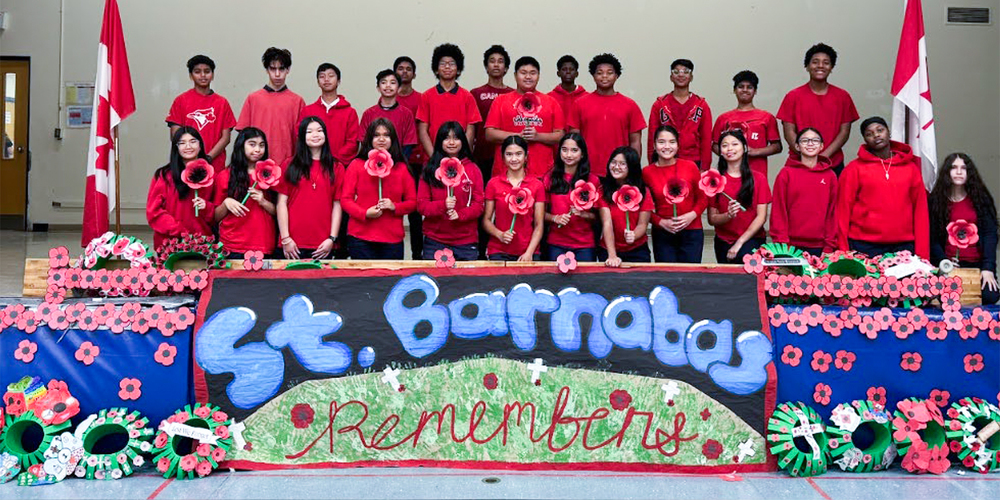 Group photo of St. Barnabas students wearing red and holding poppies in front of a St. Barnabas Remembers banners, flanked by two Canada flags