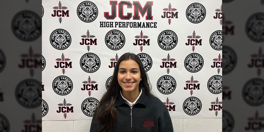 Photo of Cecilia standing in front of a wall papered with the JCM High Performance and Wolfpack logo