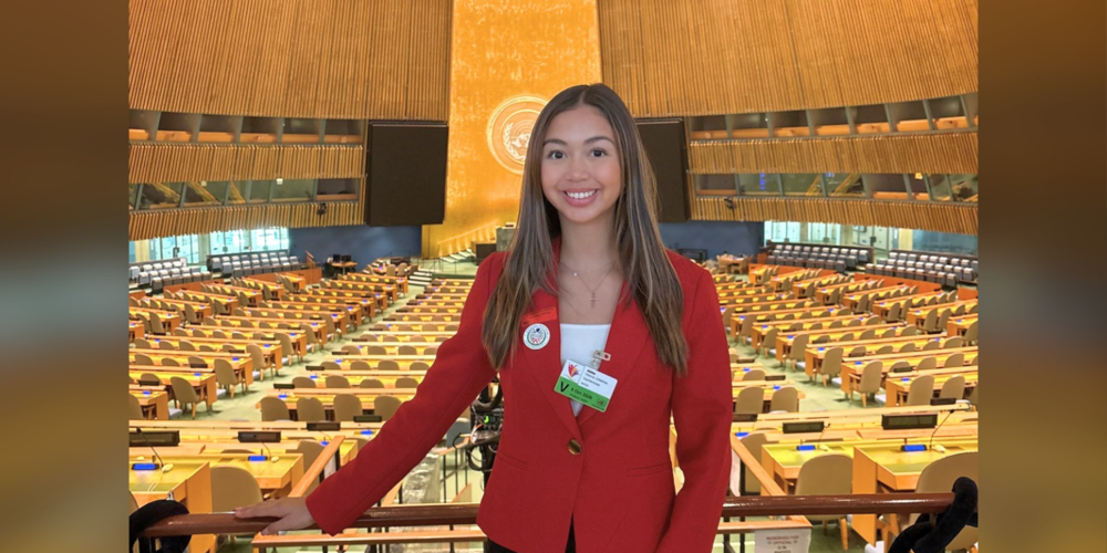 Person standing in a large parliamentary chamber with rows of yellow seats and desks, warm lighting overhead, and a circular emblem on the back wall, wearing a red blazer and holding a badge.