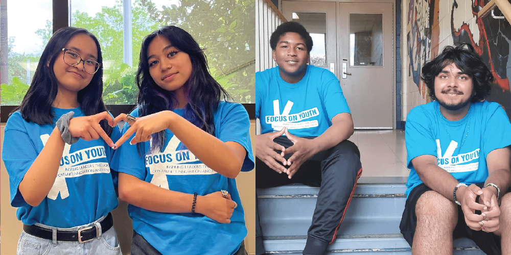 Two youths wearing matching blue ‘Focus on Youth’ T‑shirts forming a heart shape with their hands, next to two other youths in the same T‑shirts sitting on school steps.