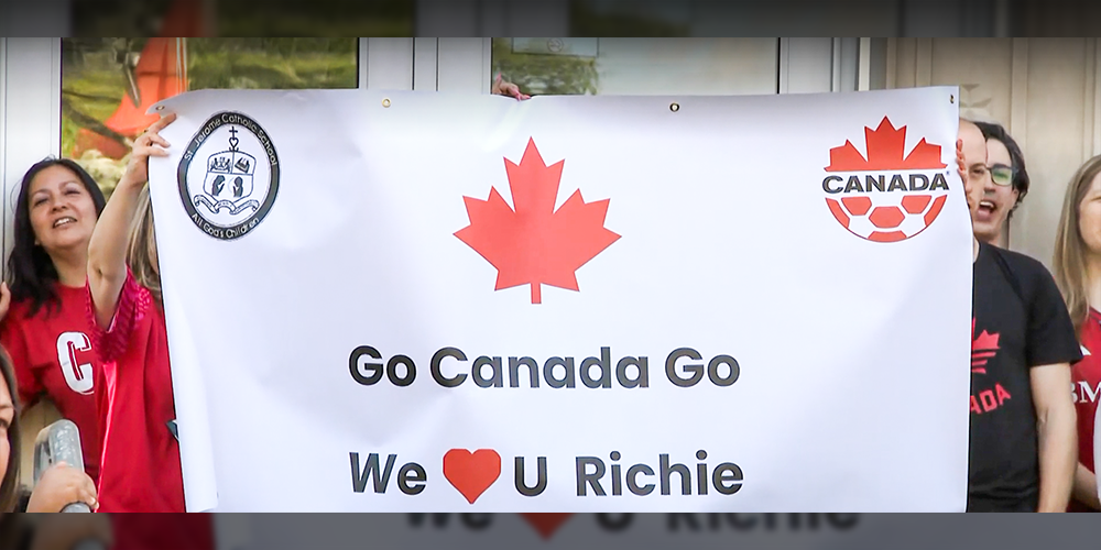 Photo of school community holding up a white banner that has the Canada red maple leaf on it, as well as the words "Go Canada Go We Heart You Richie"