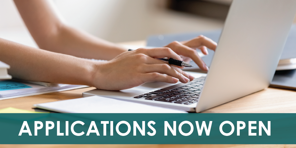 Close-up of a person’s hands typing on a laptop while holding a pen at a desk, with a teal banner across the bottom displaying the text “APPLICATIONS NOW OPEN.”