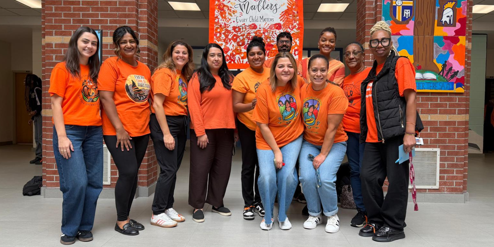 Group of people in orange shirts posing for a photo outside a brick building hallway, standing in front of a red-orange banner and a colorful mural on the wall.