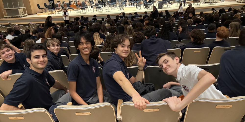 Students in a lecture hall sit in rows of gray seats, smiling and posing for the camera while others sit in the background, with a stage and orchestra instruments set up at the far end.