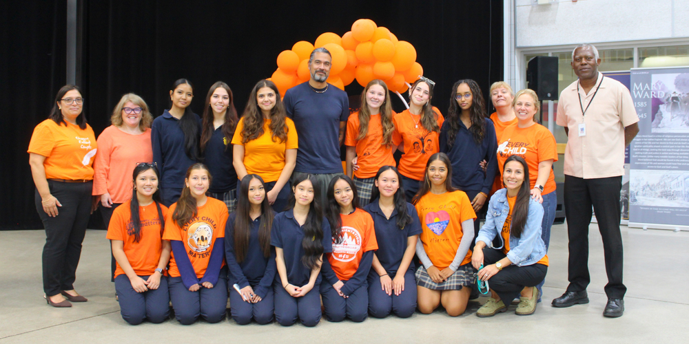 Group photo of event participants standing in front of a backdrop with a large orange balloon cluster, with several people wearing orange shirts and a few in dark clothing.