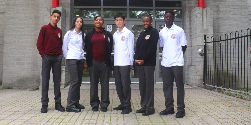 A group of five secondary school students in school uniform stand outside a school entrance, posed on a paved walkway in front of the building. The students wear burgundy, white, and navy uniform tops with grey pants