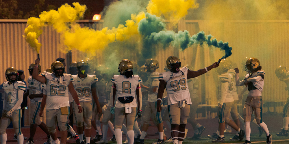 Photo of Chaminade football student athletes in full gear on the court, setting out colorful plumes of smoke in the Chaminade colours of yellow and green