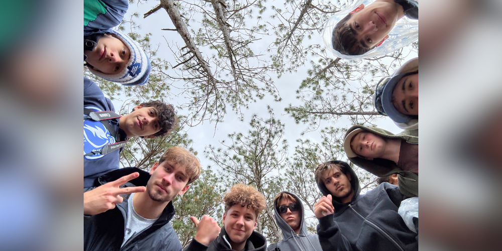 Low-angle selfie-style group photo of several people outdoors in winter, standing in a circle and looking up at the camera beneath leafless trees and a cloudy sky.