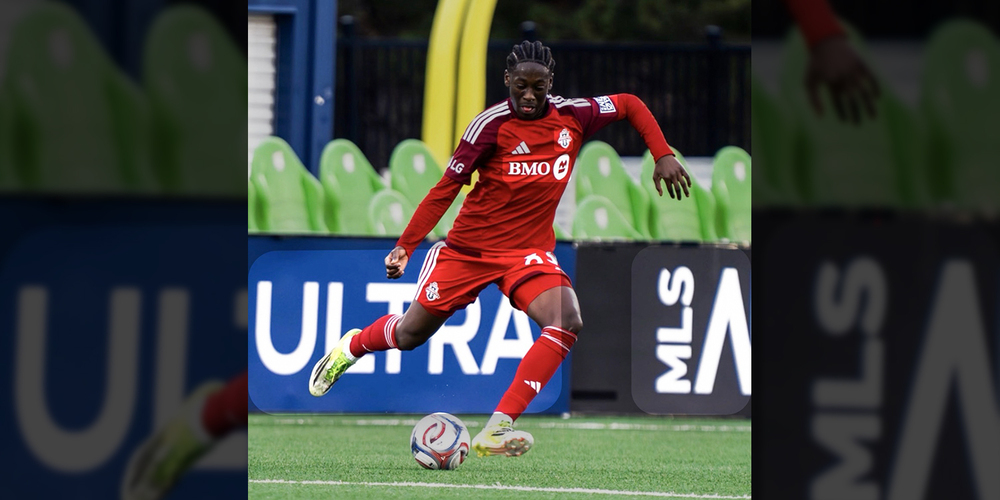 Edwin, a Toronto FC player in a red kit runs toward a soccer ball on the field, poised to kick, with stadium seats and MLS signage behind him.