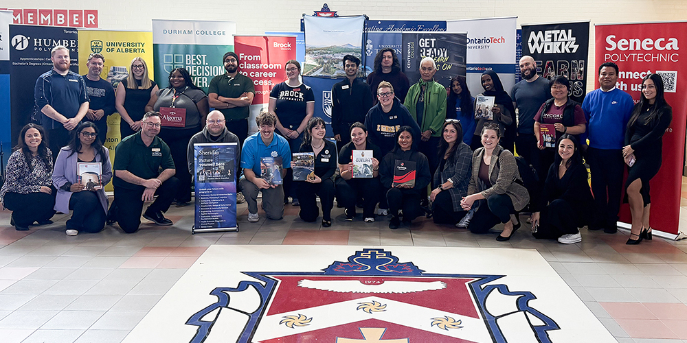 Group photo of presenters at the fair with their organization's banners behind them, posing in the FHC school building, with the FHC school crest emblazoned on the floor in front of them
