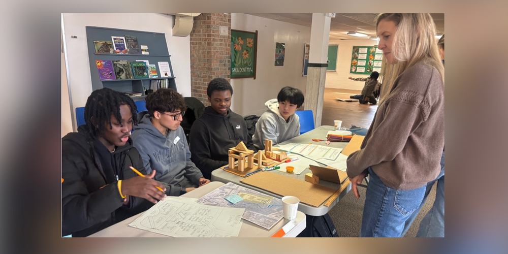 A teacher standing while students are at a desk working. 