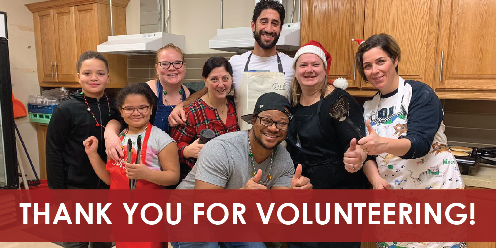 A group of eight people standing together in a kitchen setting, some wearing aprons and festive accessories, posing and giving thumbs‑up gestures. A red banner across the bottom displays the text ‘THANK YOU FOR VOLUNTEERING!’