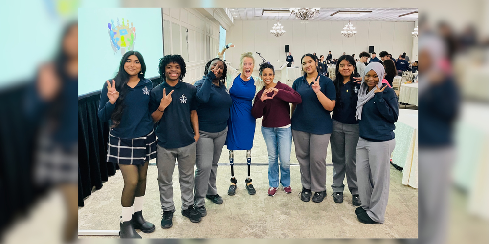 A group of eight students and two adult presenters stand together in a banquet hall, smiling and making hand-sign gestures toward the camera. The students are wearing school uniforms, and one of the presenters, dressed in a blue dress, has visible prosthetic legs. Round tables and a large presentation screen are visible in the background.