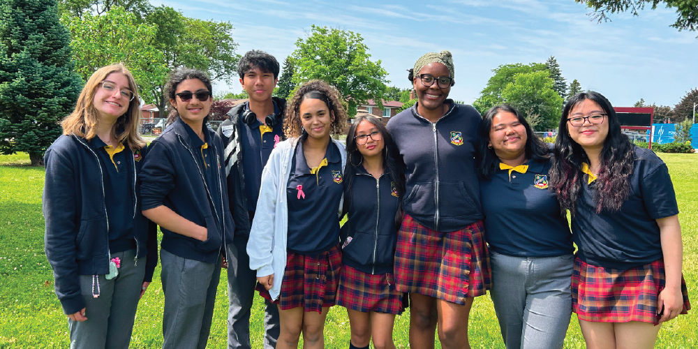 A group of students wearing school uniforms stand together outdoors on a grassy field, with trees and school buildings visible in the background on a sunny day.
