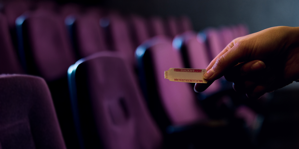 Photo of a person holding a ticket in their hand in front of a row of purple theatre seats