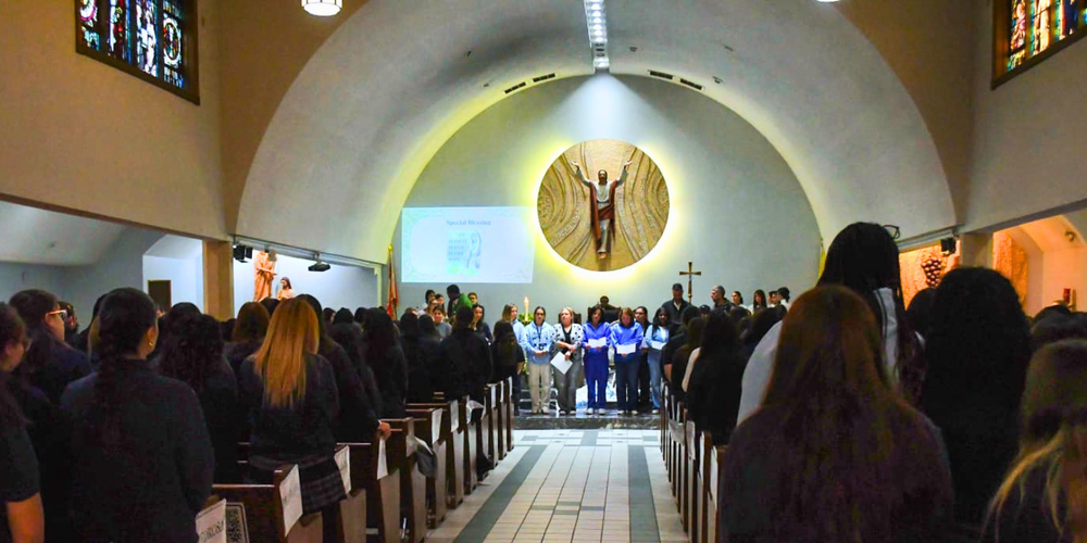 Inside a church, people stand in rows facing a choir on the altar beneath stained-glass windows and a large illuminated wall with a central statue.