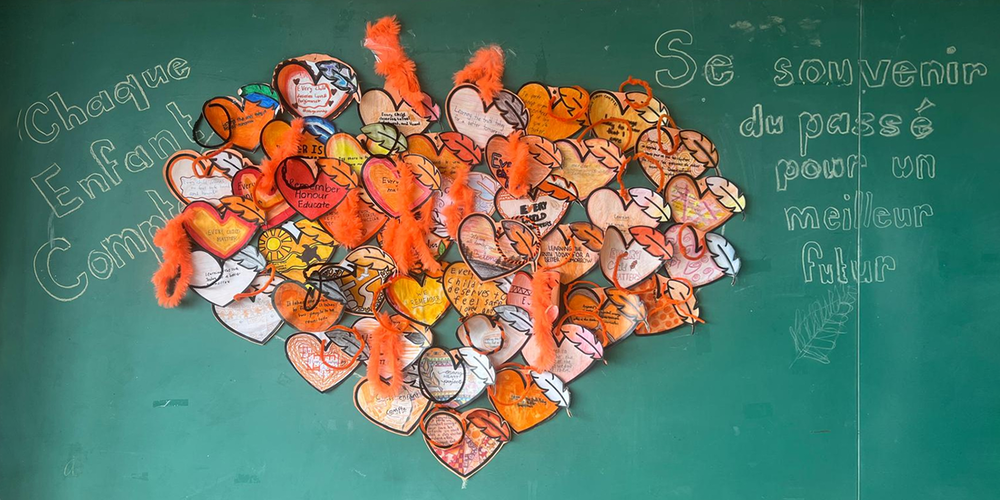 A colorful heart collage of paper hearts and orange feathers decorates a green classroom chalkboard, with French writing about a better future.