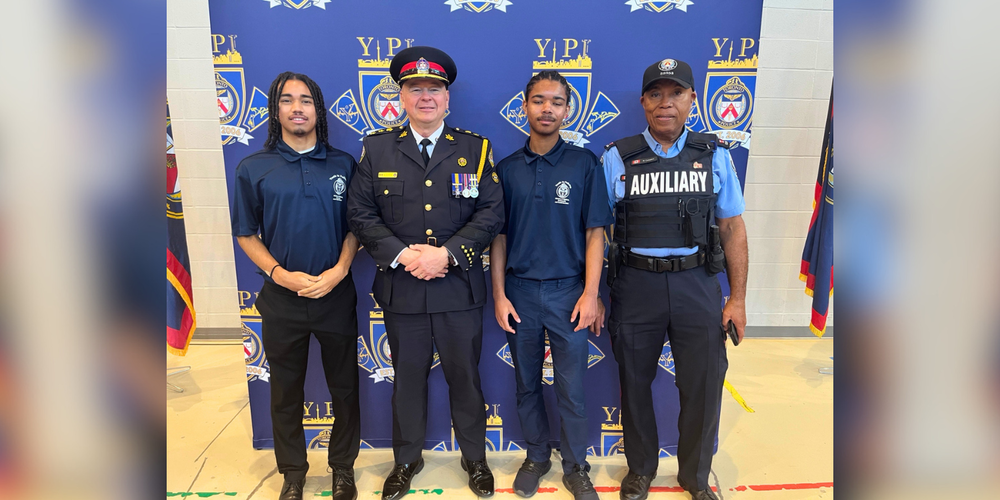 Four people pose for a photo in front of a Toronto Police Service Youth in Policing Initiative backdrop, including two youth participants, a uniformed police officer and an auxiliary officer.