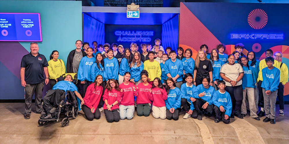 Group photo of Redmond students posing in blue, red and yellow team shirts along with Redmond staff in front of a neon "Challenge Accepted" sign at BRKFREE