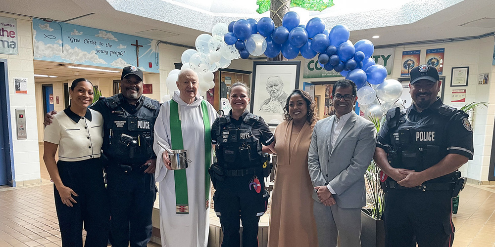 Group photo of the priest, the Chaplain, police officers, Principal Navaratnasingham and Superintendent Peterson in front of a drawing of St. John Paul II
