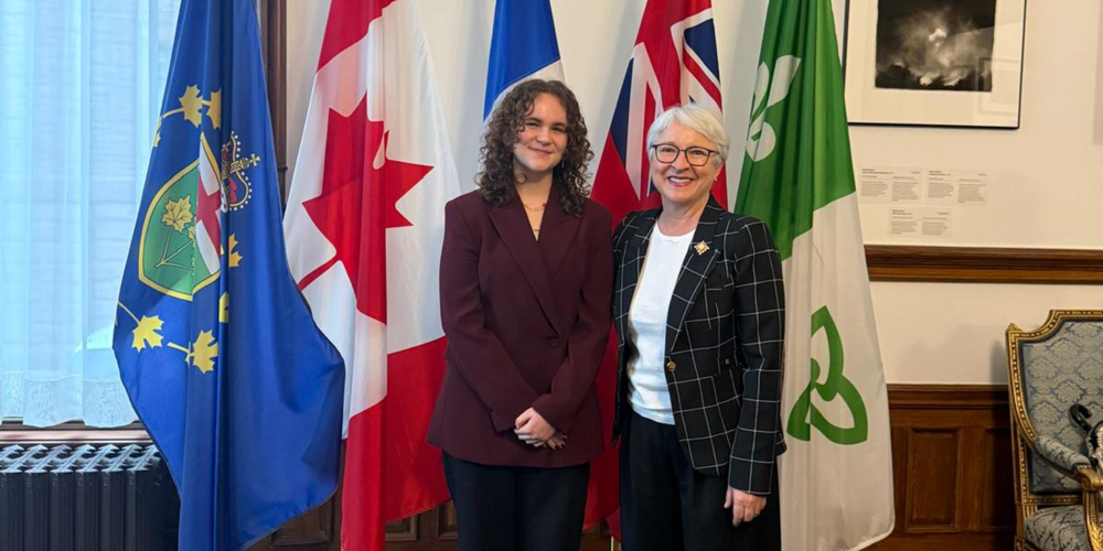 2 women standing in front of flags, dressed in business attire. 